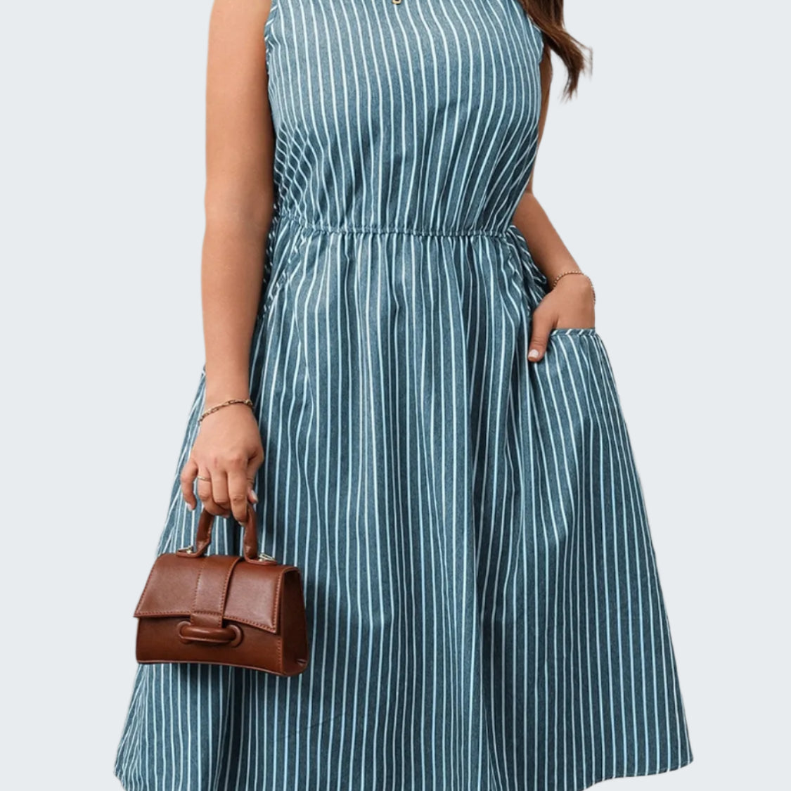 A model wearing a plus size striped round neck sleeveless dress in blue and white, with a brown belt, holding a brown bag.
