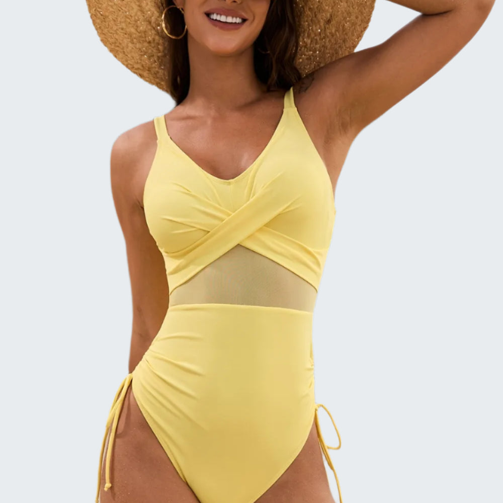 A woman at the beach wearing a yellow one-piece swimsuit with a v-neck and drawstrings. She is also wearing a wide-brimmed straw hat.