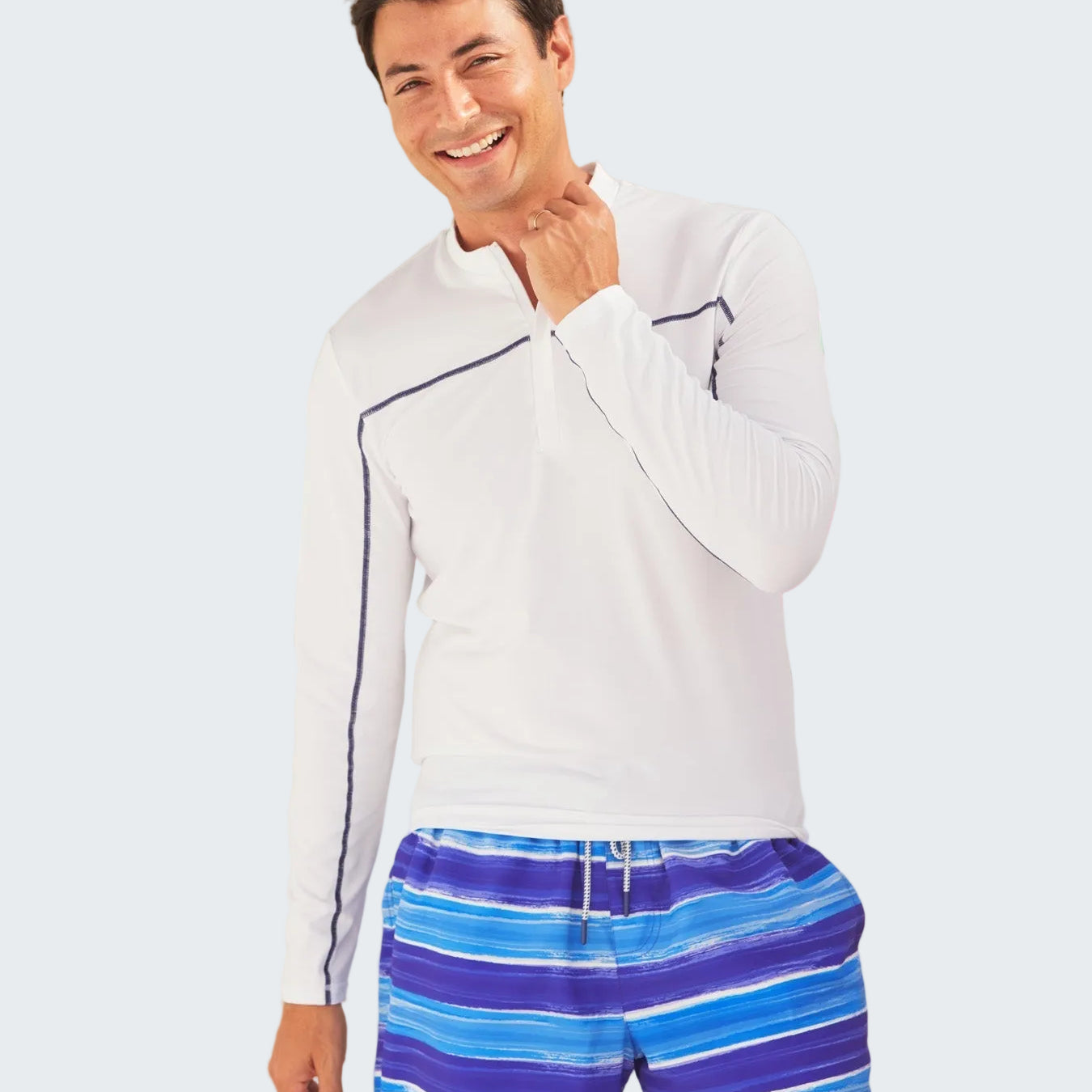 Man wearing a white long-sleeve shirt and blue striped shorts standing in front of a polka dot surfboard.