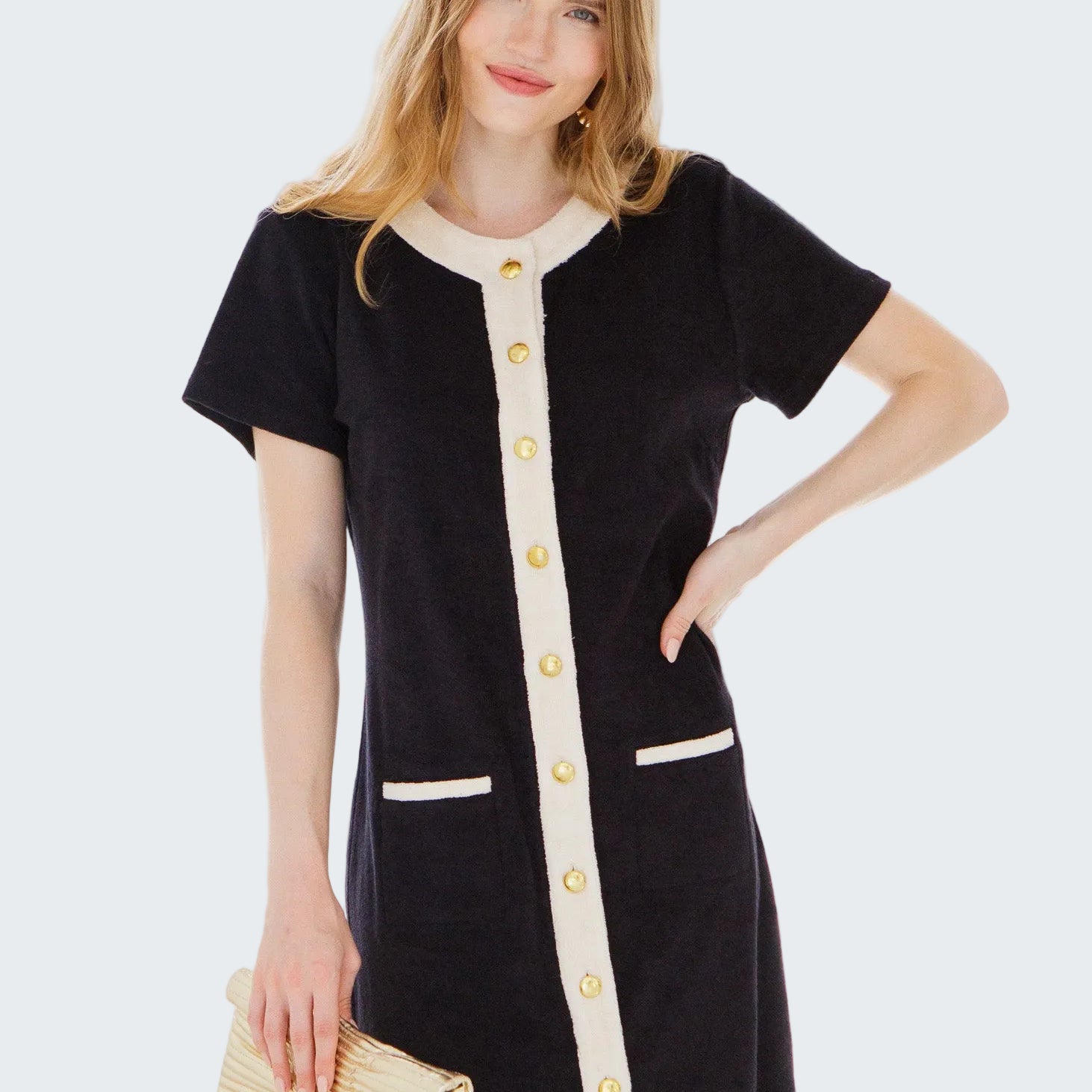 Woman wearing a black dress with white buttons and trim, holding a beige clutch in front of a window.