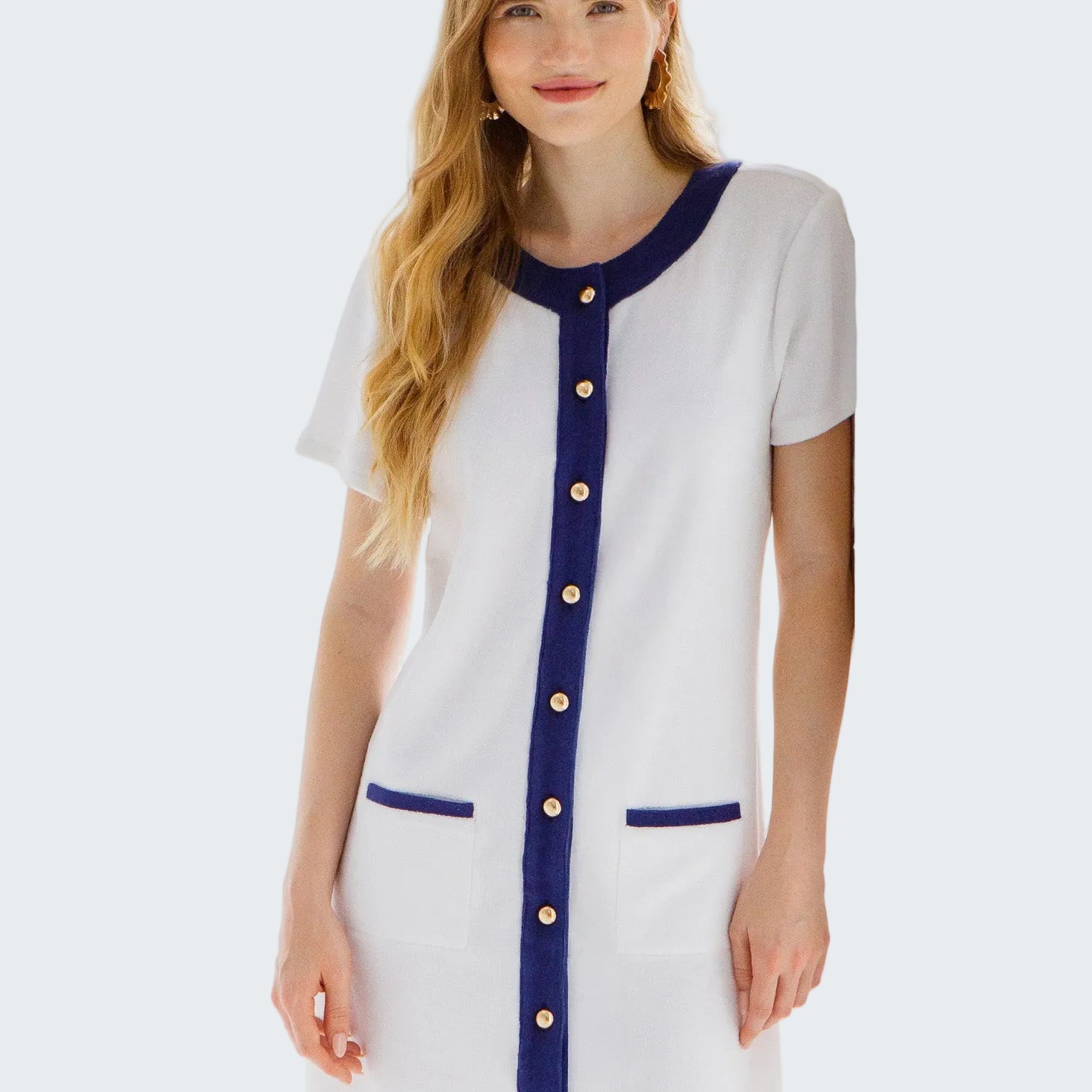 Woman wearing a white dress with blue accents and gold buttons in a bright room.