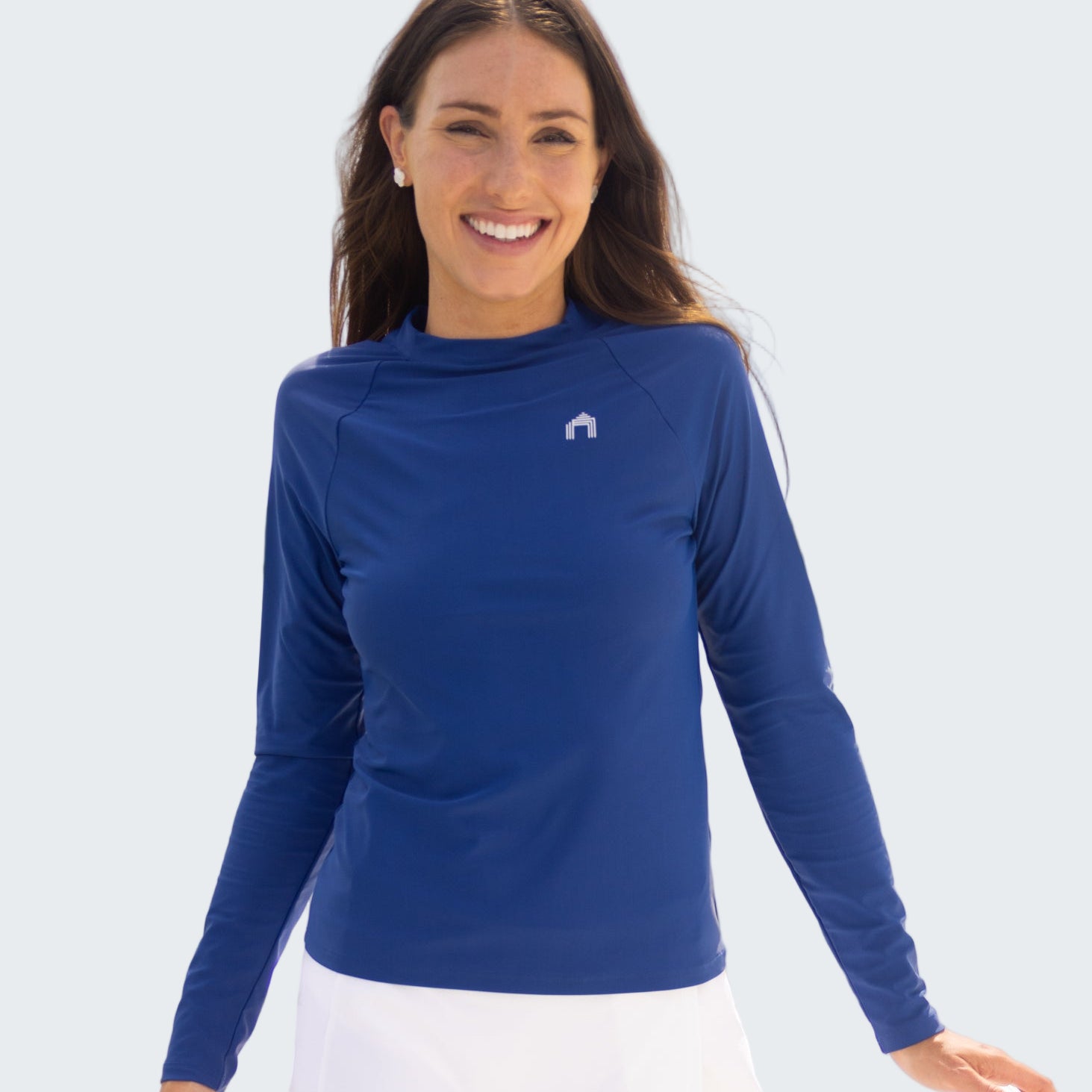 Woman wearing a blue long-sleeve shirt and white skirt standing by a body of water with sailboats in the background.