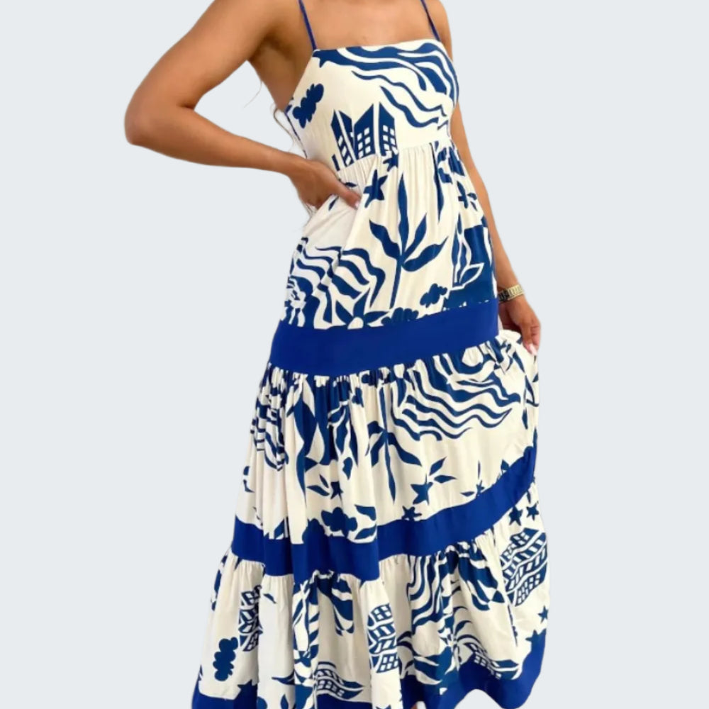 A model wearing a square neck, floral printed maxi dress with a blue belt and trim, standing in a room with a white background.