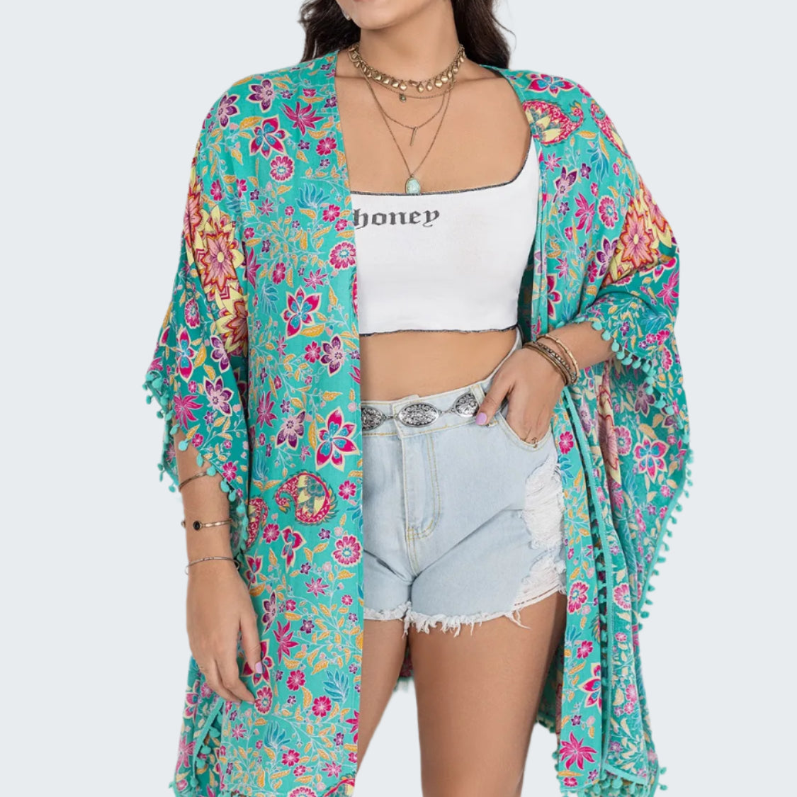 A woman wearing a multicolored floral print open front cover-up with tassel detailing, paired with a white top and blue denim shorts.