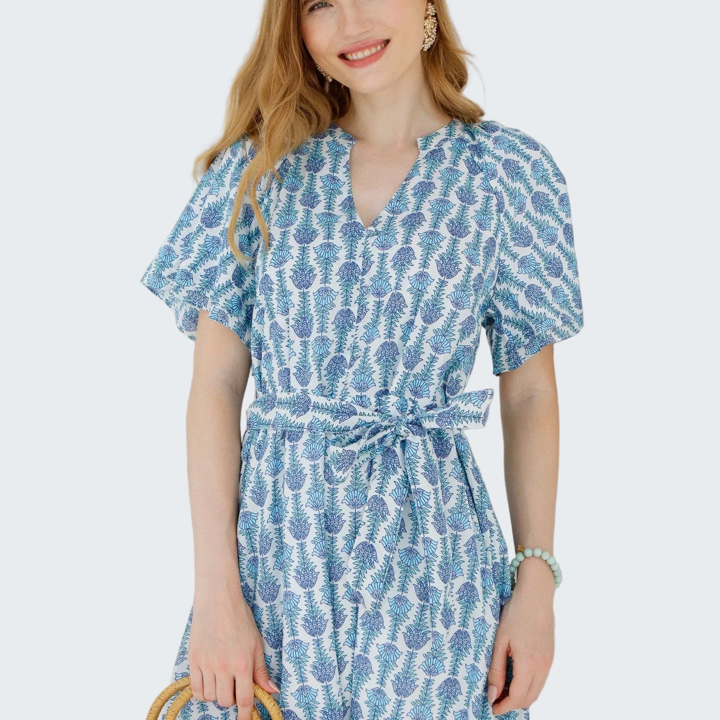 Woman wearing a blue patterned dress holding a straw bag against a white wall.