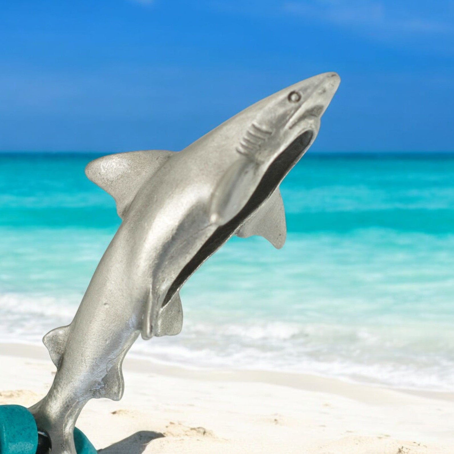 Metallic shark figurine on a sandy beach with turquoise water and blue sky in the background