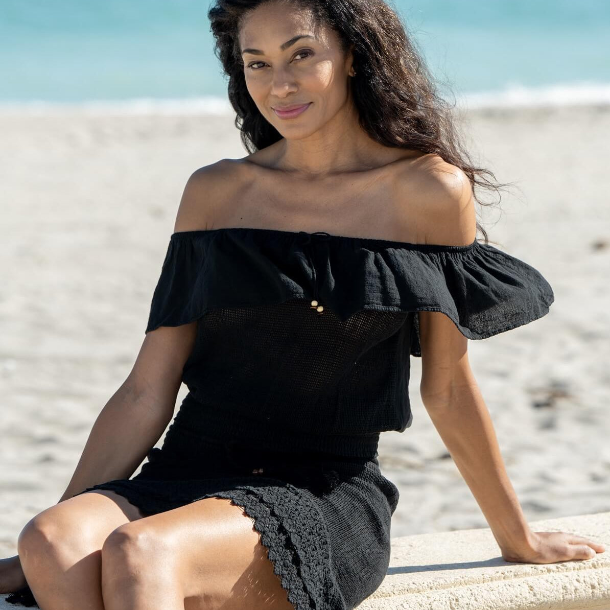 Woman in a black off-shoulder dress sitting on a beach.