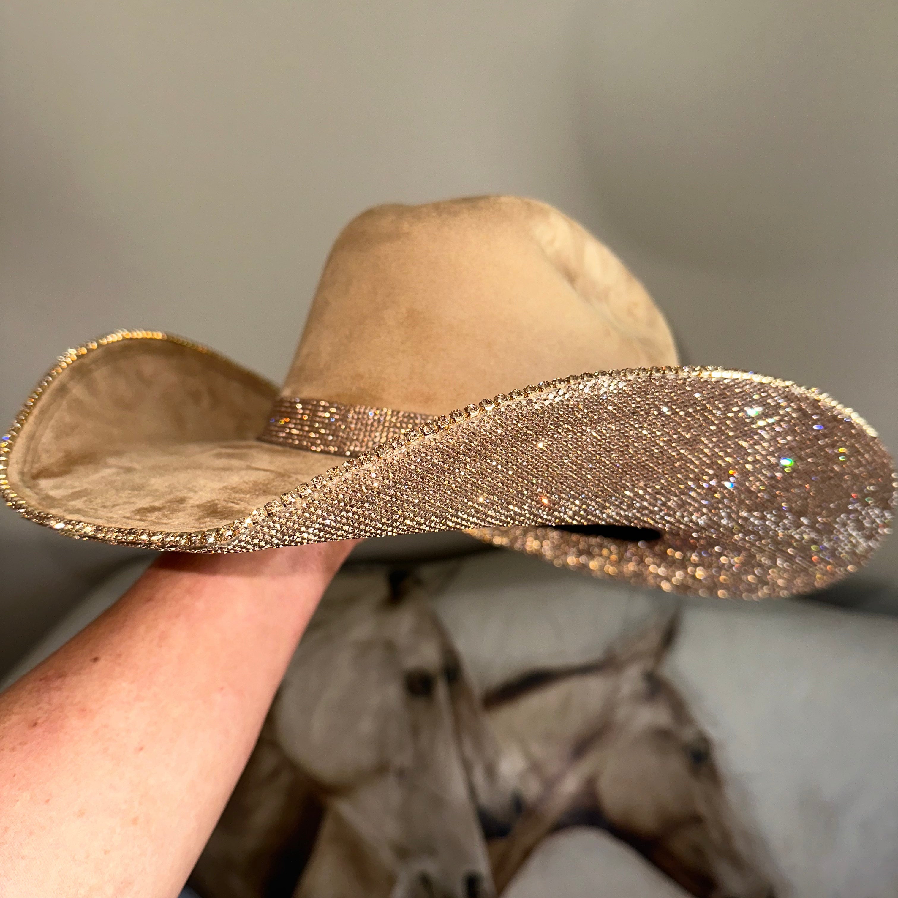 Beige cowboy hat with a glittery band held by a hand against a blurred background