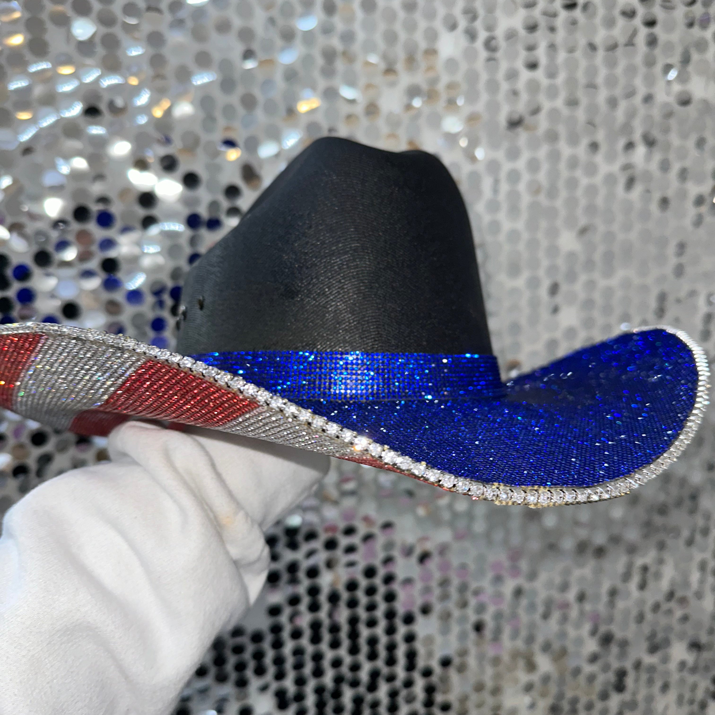 Black cowboy hat with a blue brim held by a gloved hand against a sequined background