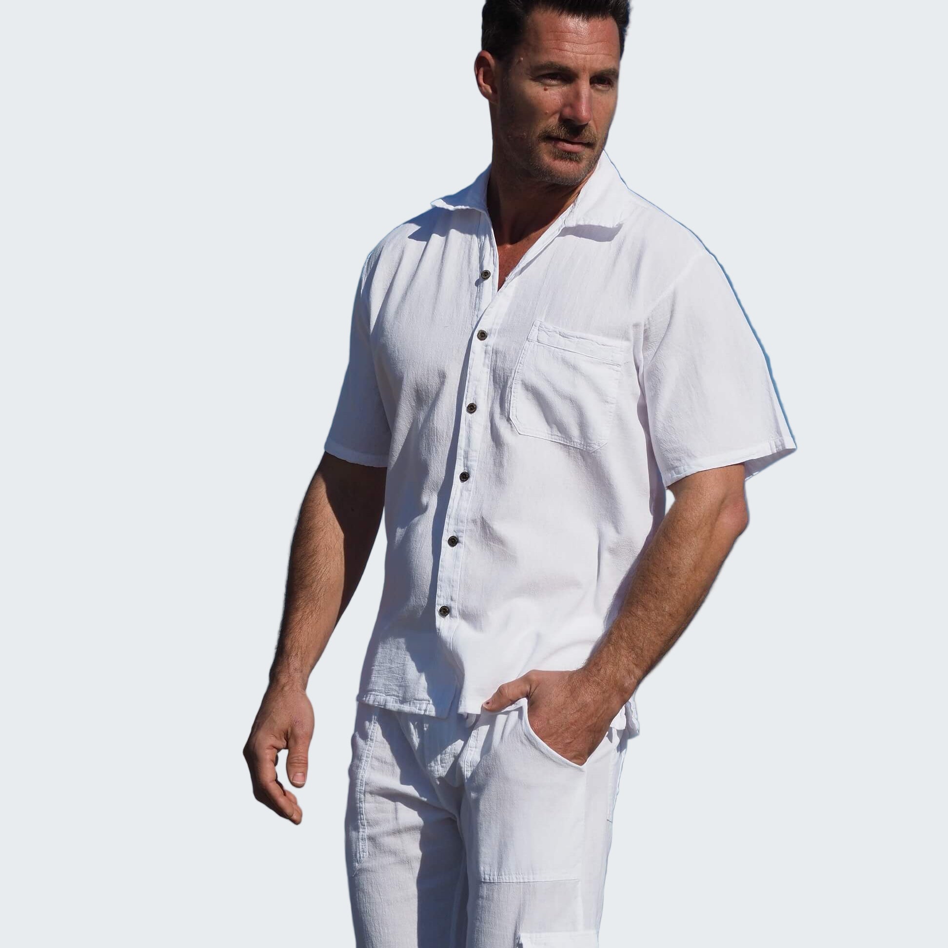 Man in a white outfit standing on a beach with palm trees and blue sky.