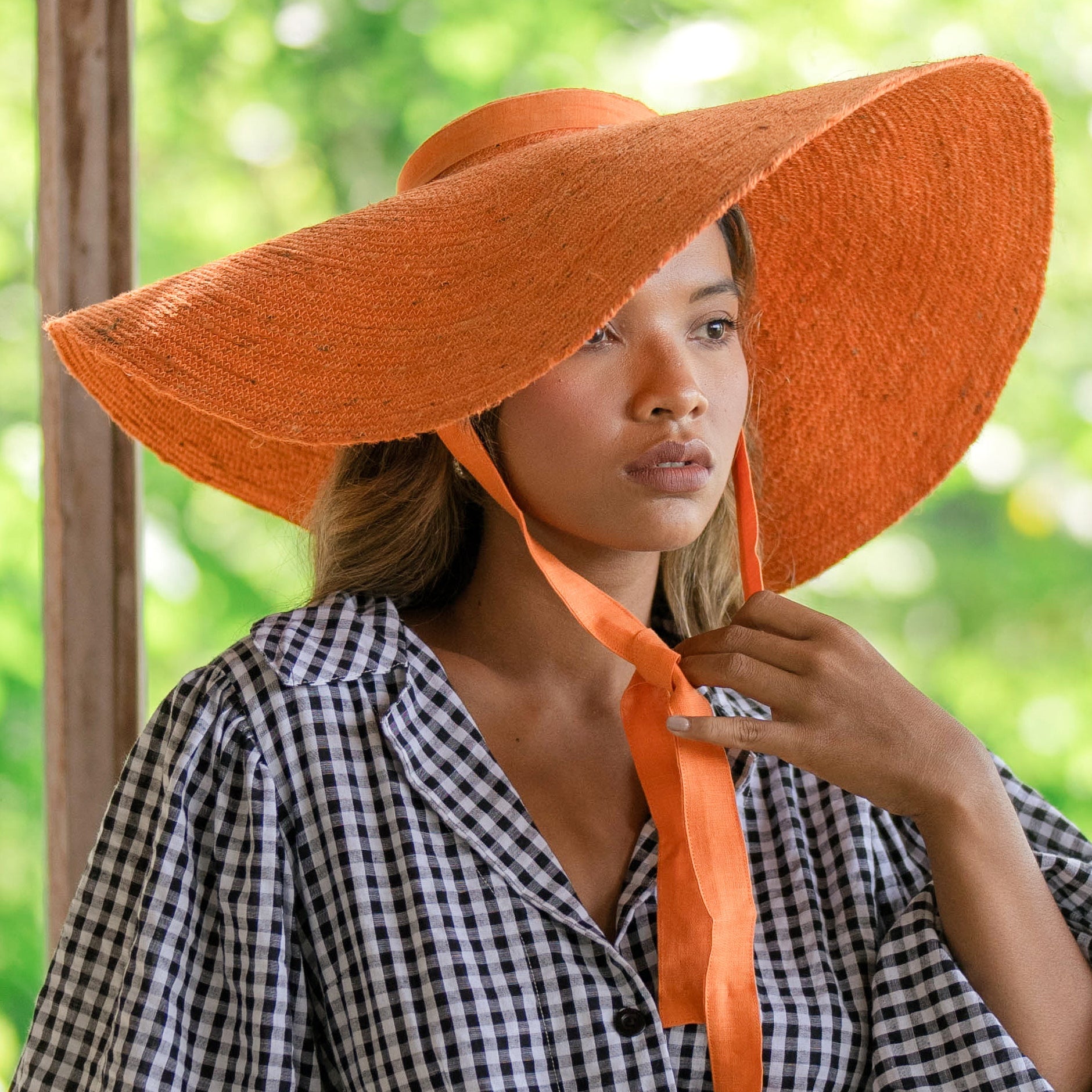 Woman wearing an orange hat with a wide brim and matching scarf, standing outdoors with greenery in the background.