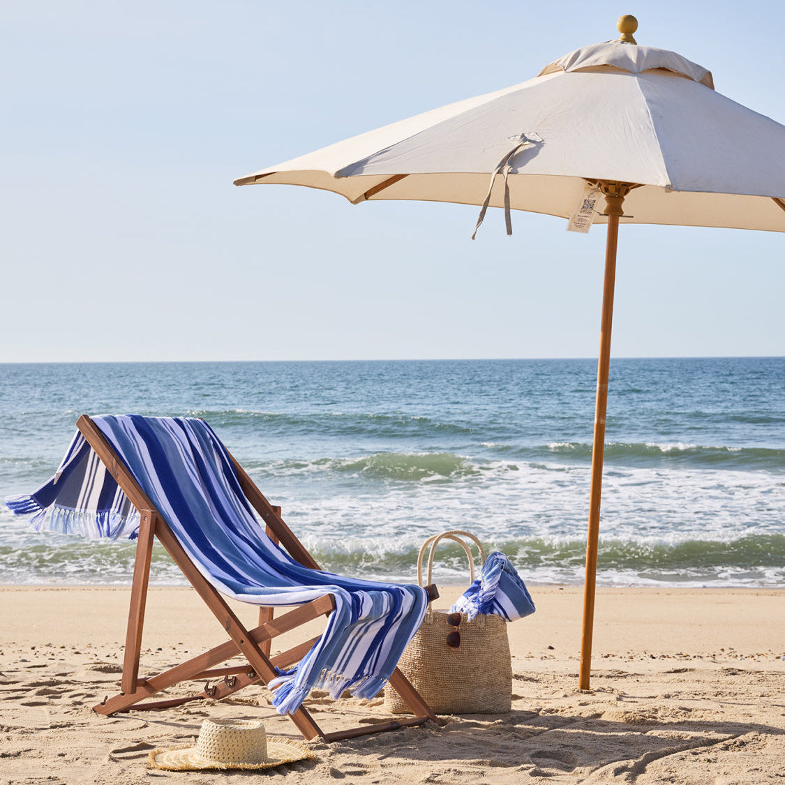 Beach chair with striped towel, umbrella, and basket on a sandy beach with ocean view.