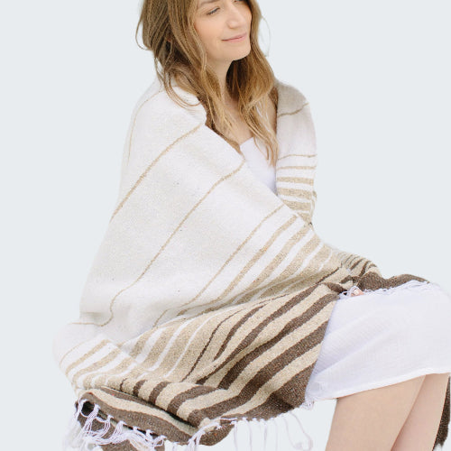 Woman sitting on a bench wrapped in a striped blanket with a neutral background