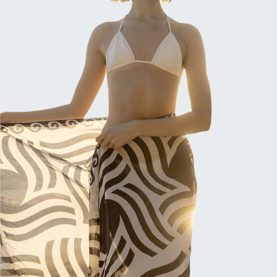 Woman in a bikini and patterned sarong standing on a beach.