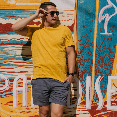 Man in yellow shirt and sunglasses holding a surfboard in front of a colorful mural.