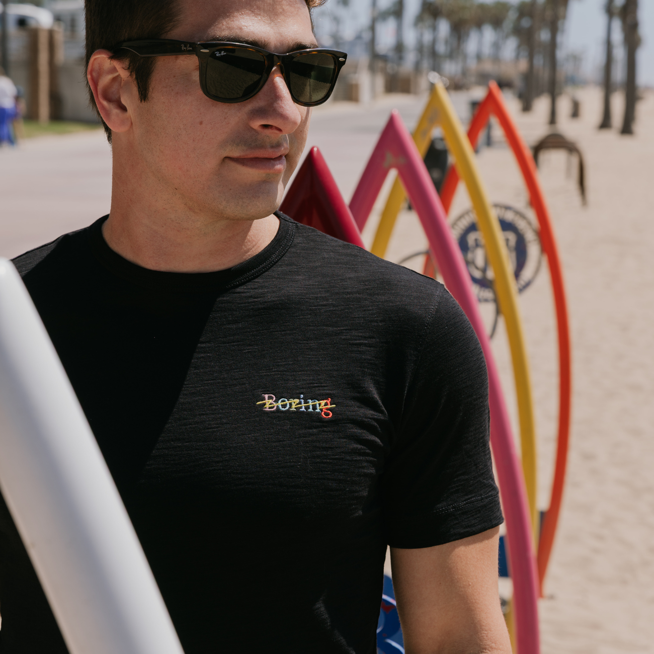 Man wearing sunglasses and a black t-shirt with a logo, standing on a beach with surfboards in the background.