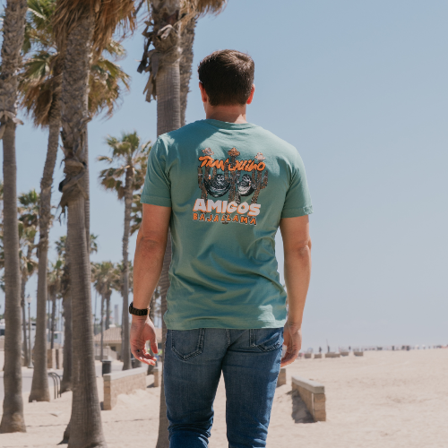 Man walking on a beach with palm trees, wearing a green t-shirt with text and graphics.