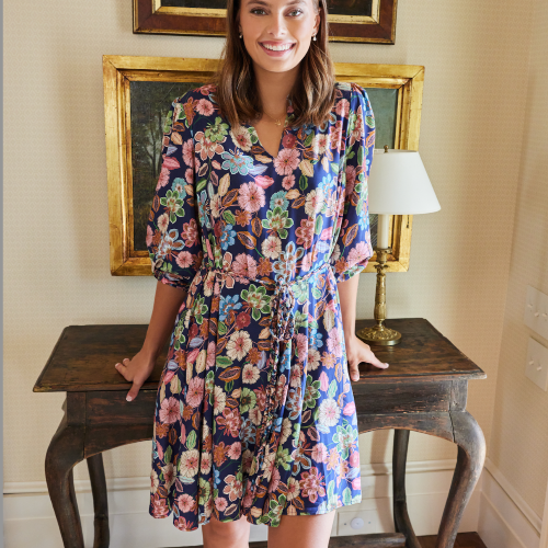 Woman in a floral dress standing in a room with wooden table and framed artwork.