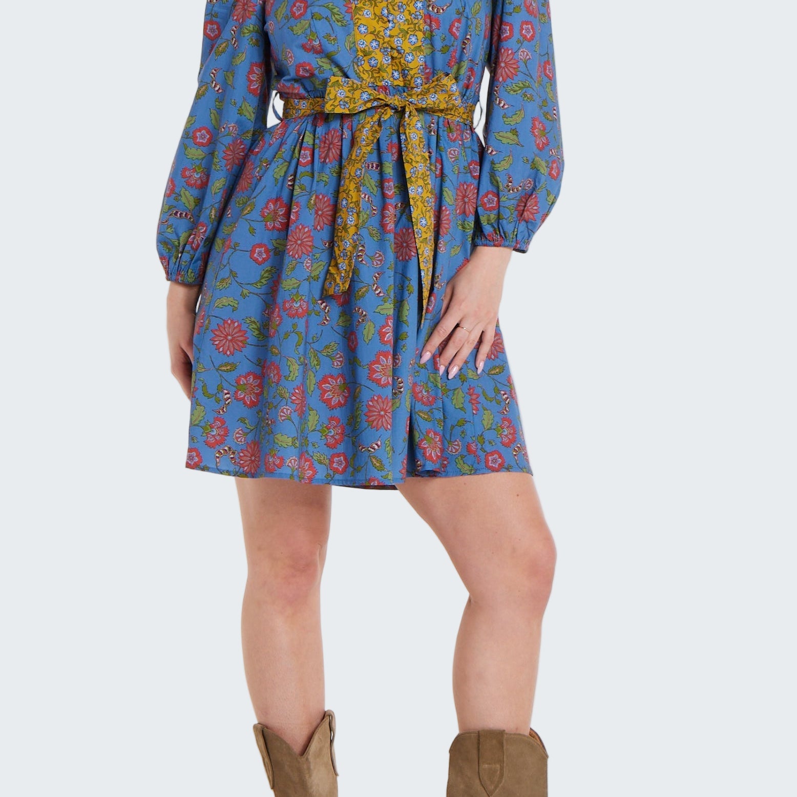 Woman wearing a patterned dress with long sleeves and a belt, paired with brown boots on a white background.