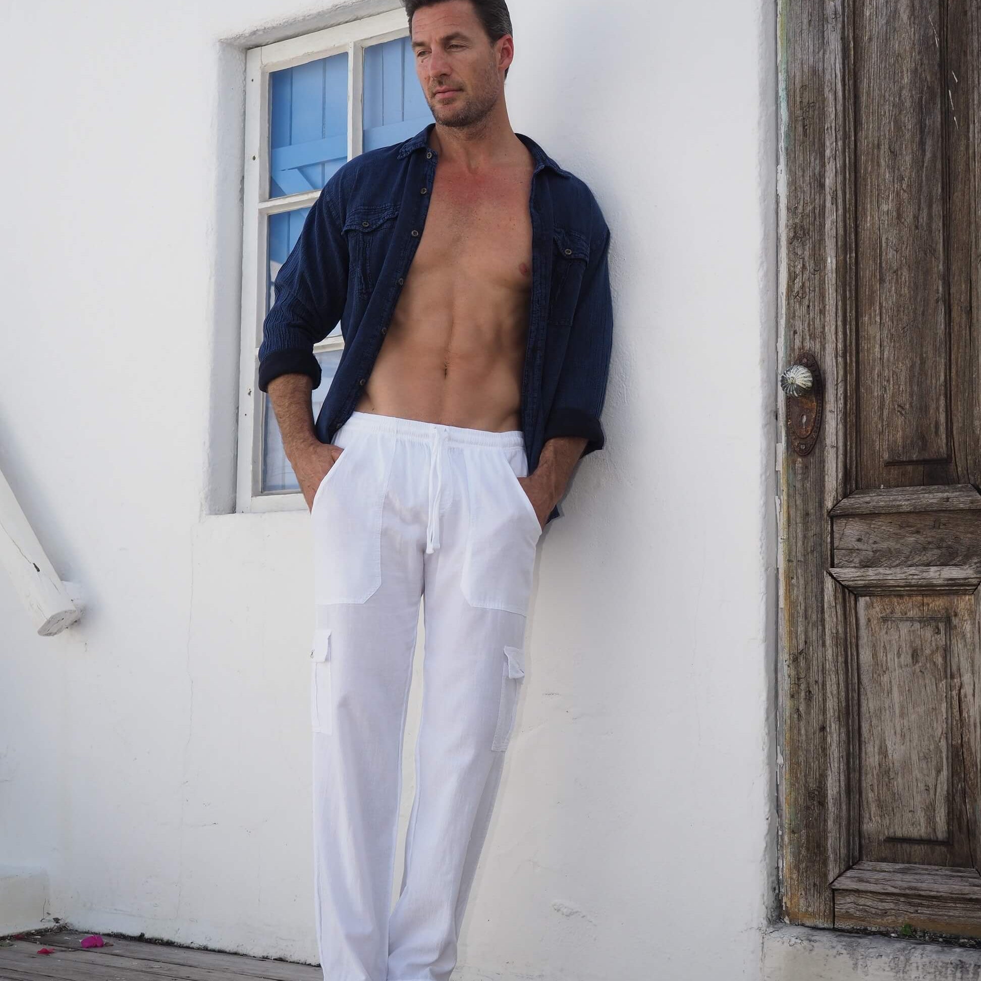 Man wearing a blue shirt and white pants leaning against a white wall with a wooden door.