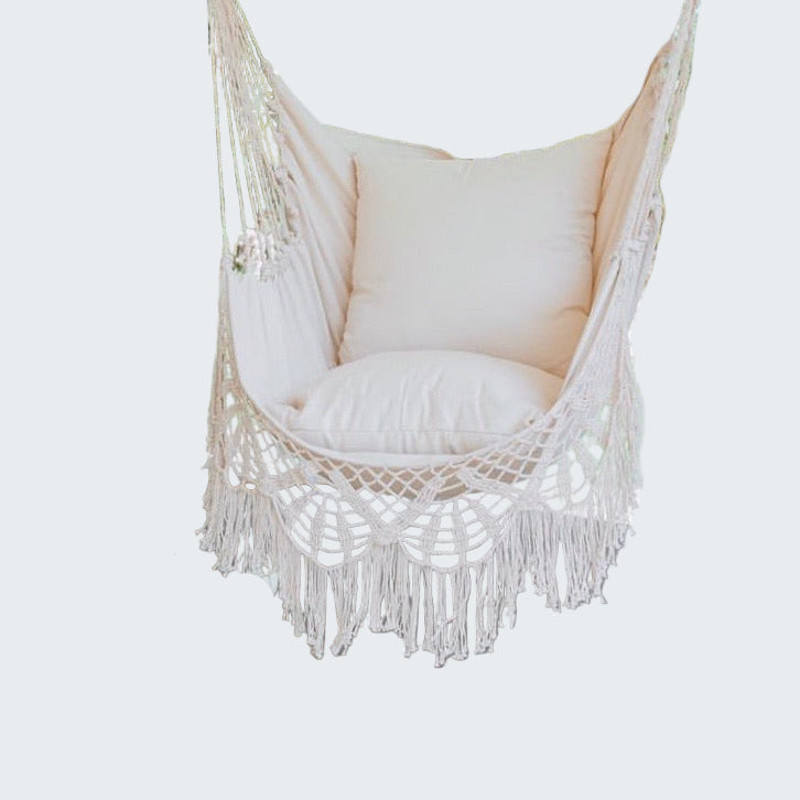 Macrame hammock chair with cushions on a wooden deck, surrounded by greenery.