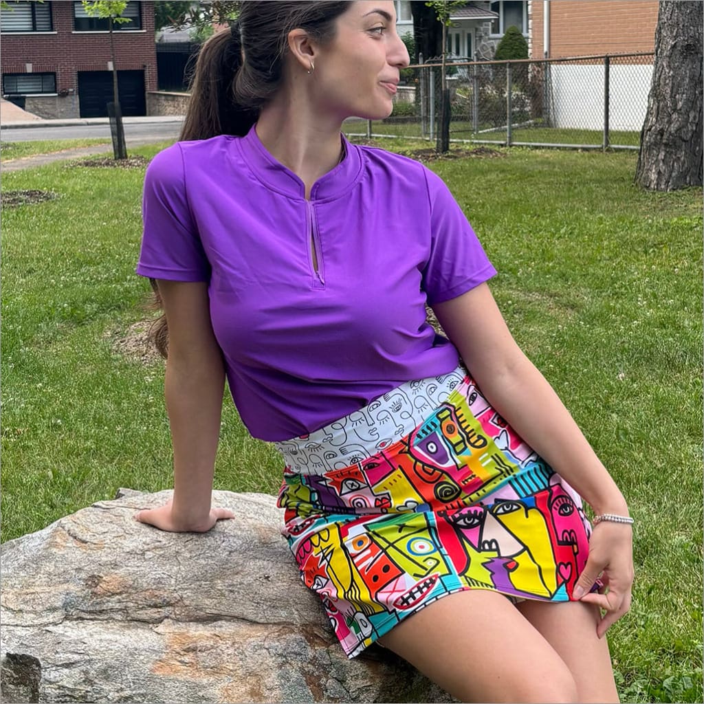 Woman in purple shirt and colorful skirt sitting on a rock outdoors.