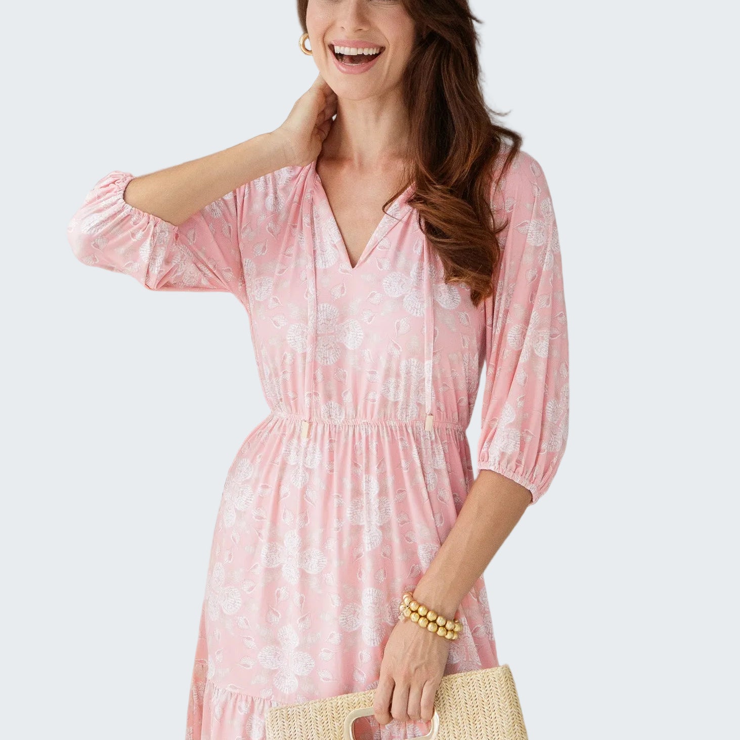 Woman wearing a pink dress with a white pattern, holding a straw bag indoors.