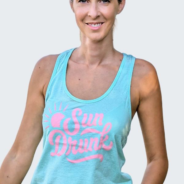 Woman wearing a light blue tank top with 'Sun Drunk' text by a body of water.