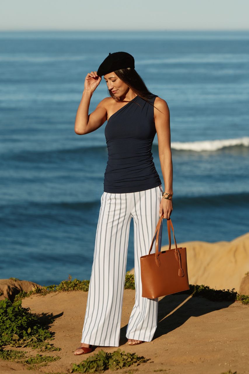 Woman standing by the ocean wearing a navy top and white striped pants, holding a brown bag.