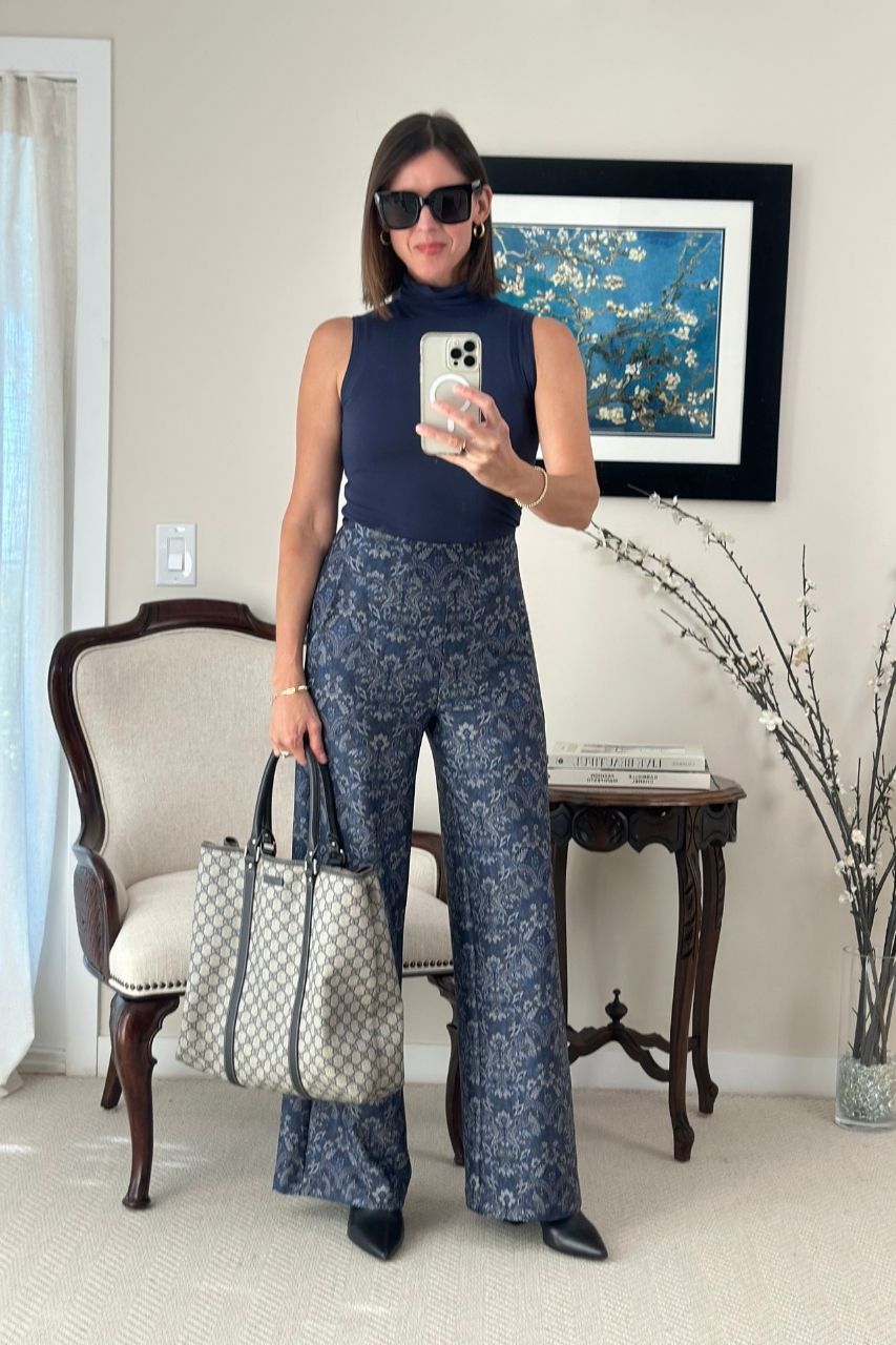 Woman taking a mirror selfie wearing a navy sleeveless top and patterned pants, holding a handbag.