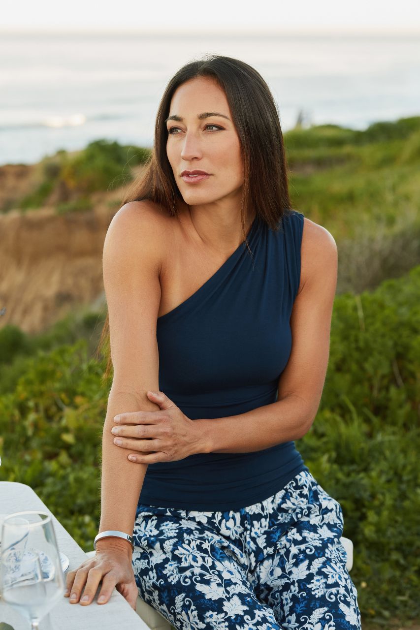 Woman in a blue top and floral pants sitting outdoors with greenery in the background