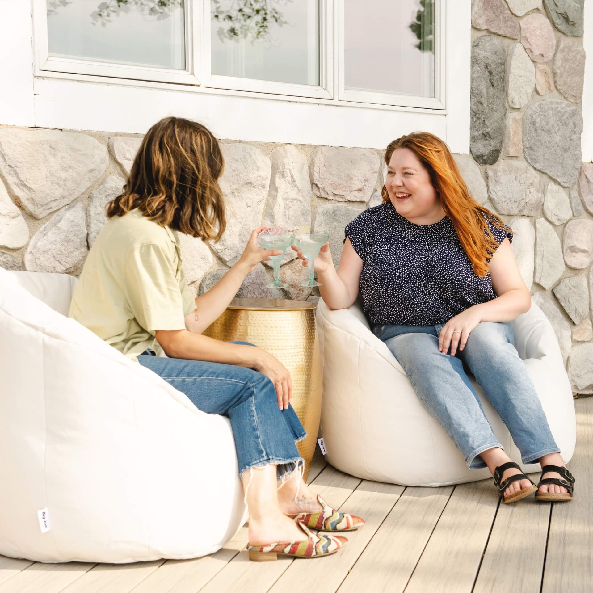 Two women sitting on bean bags in a casual outdoor setting with a stone wall background.