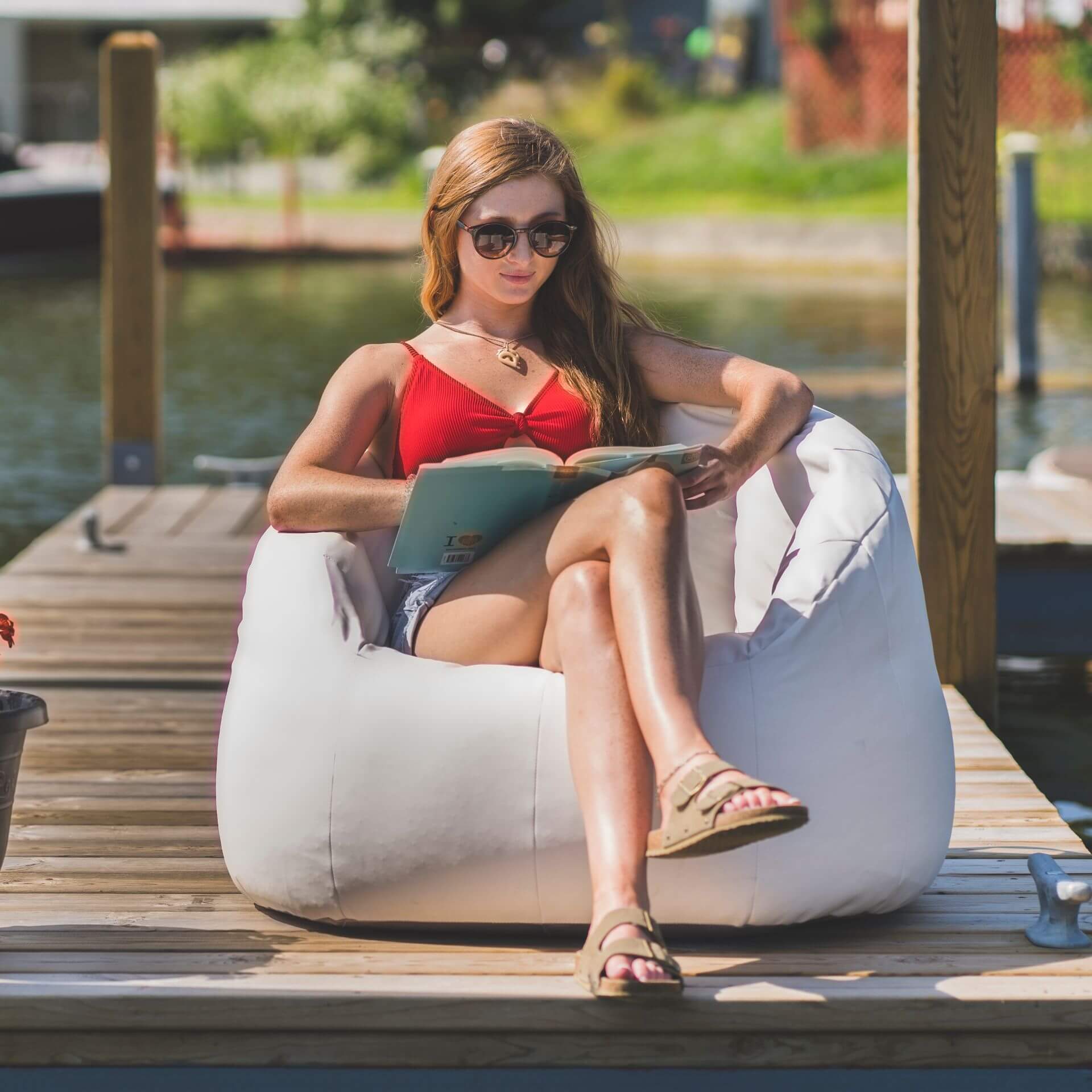 Woman reading a book on a white bean bag chair by a waterfront.