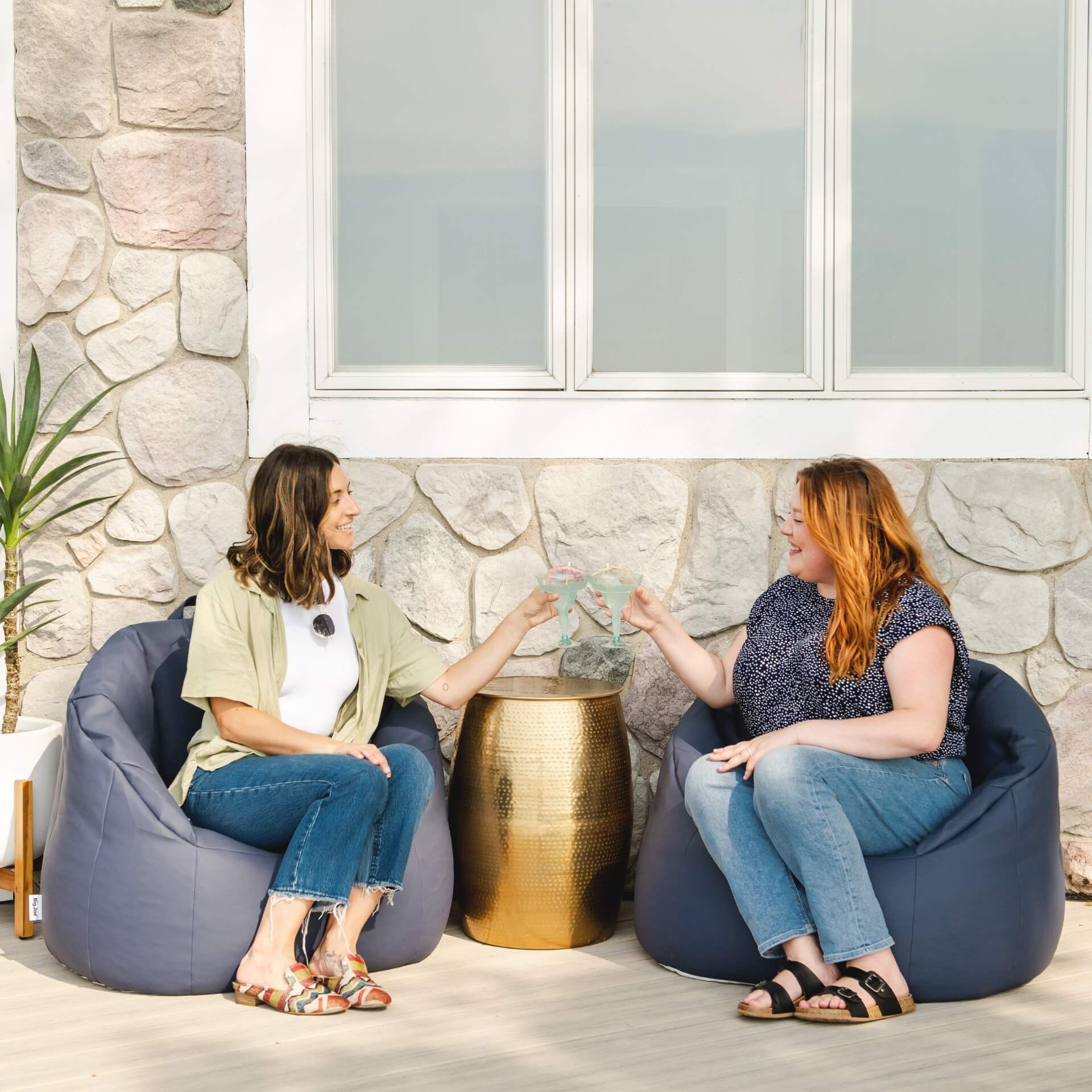 Two women sitting on bean bags in a casual setting with a stone wall and window in the background.