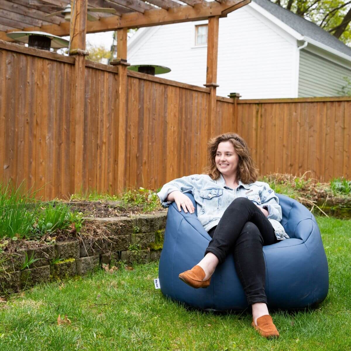 Woman sitting on a blue bean bag chair in a backyard with a wooden fence and house in the background.