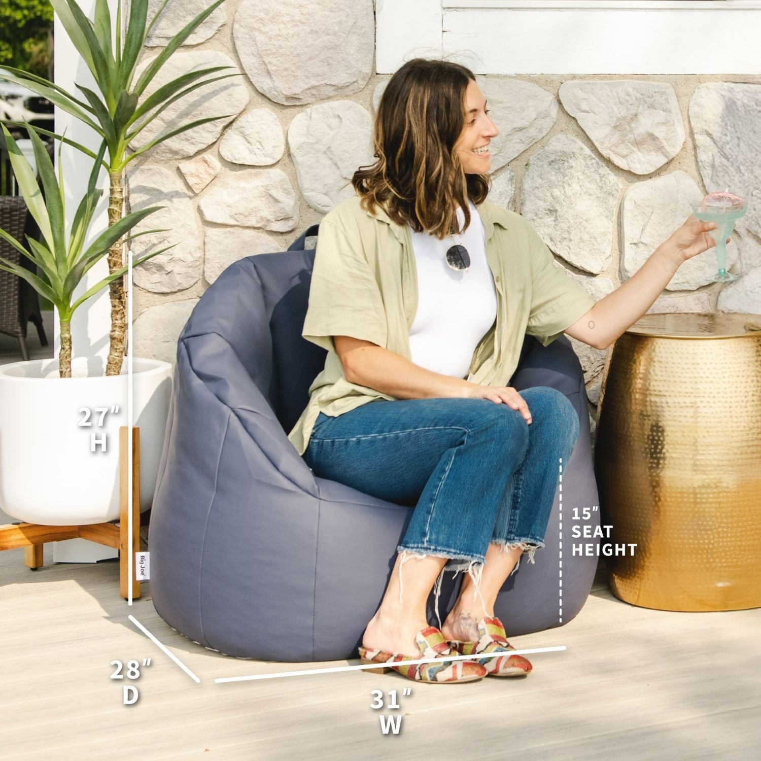 Woman sitting on a gray bean bag chair with measurements displayed, against a stone wall.