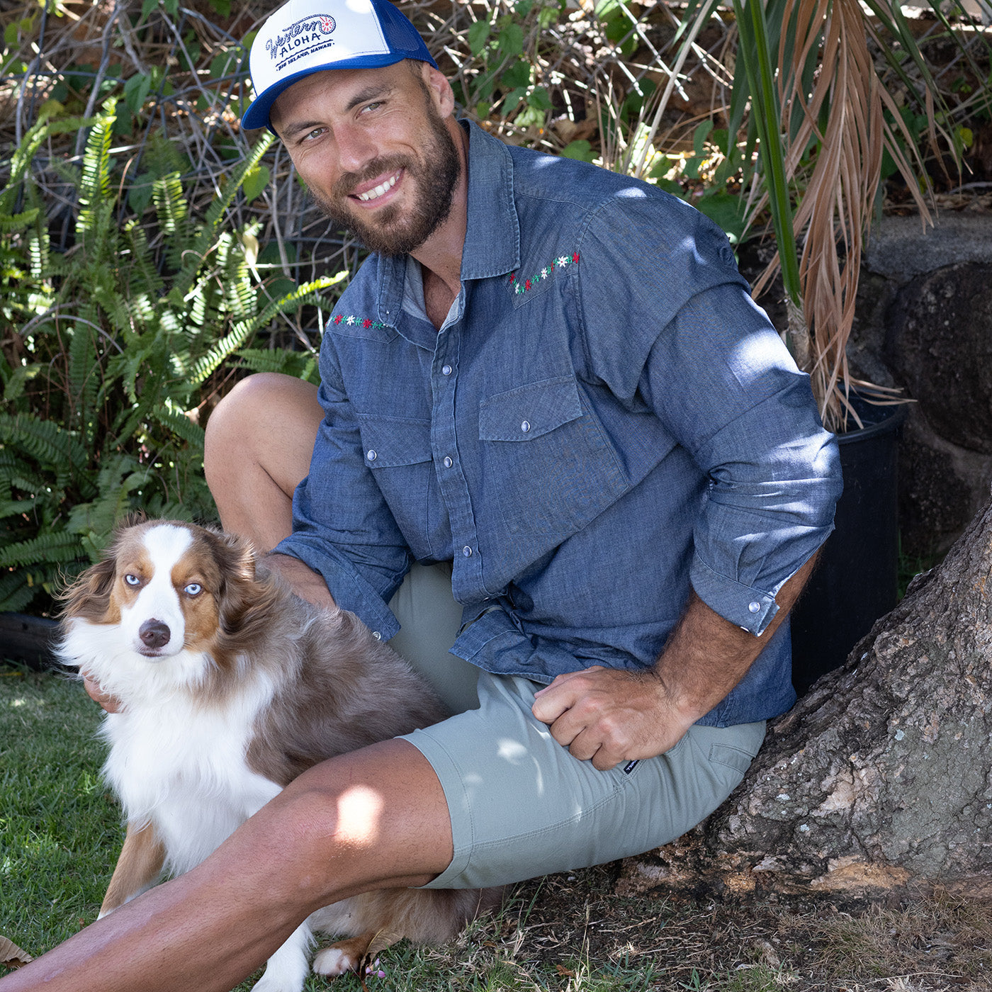 Man sitting outdoors with a dog, wearing a blue shirt and cap.