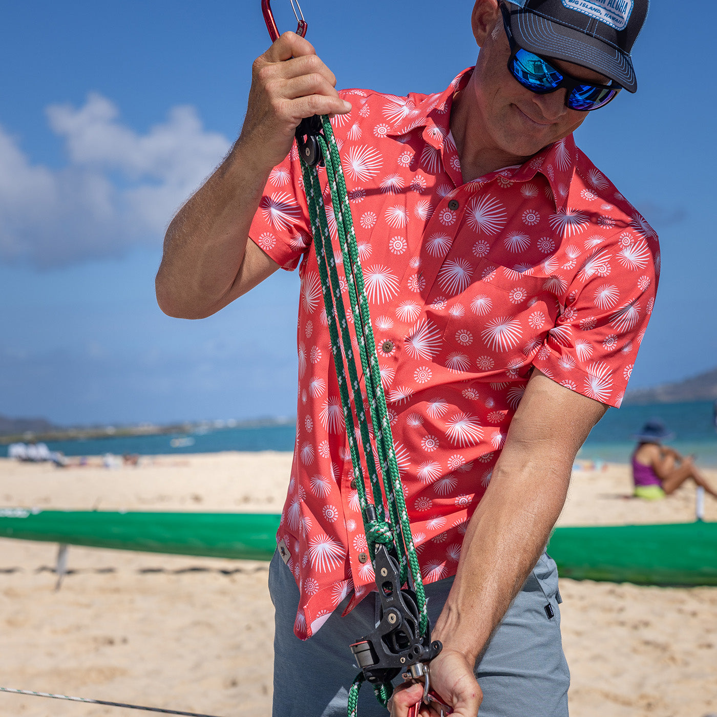 Person on a beach adjusting equipment, wearing a red patterned shirt and sunglasses.