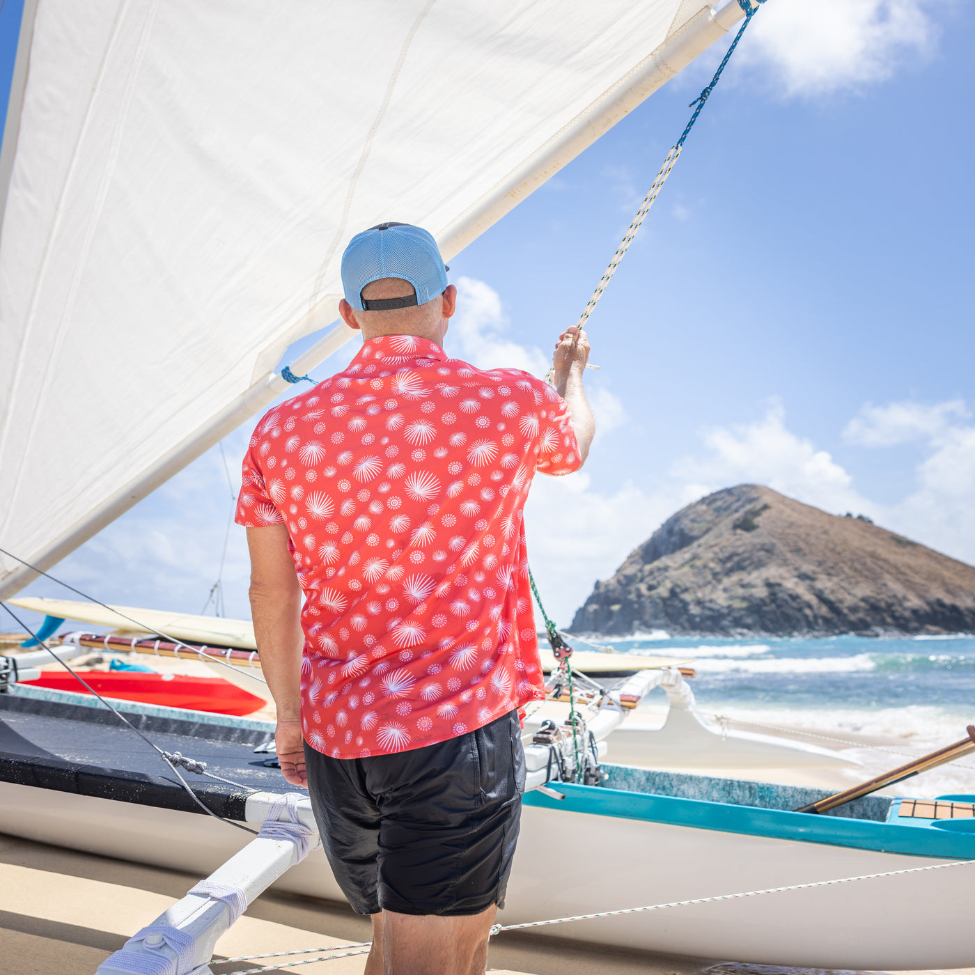 Man on a sailboat wearing a red patterned shirt and blue cap, with a scenic background of mountains and water.