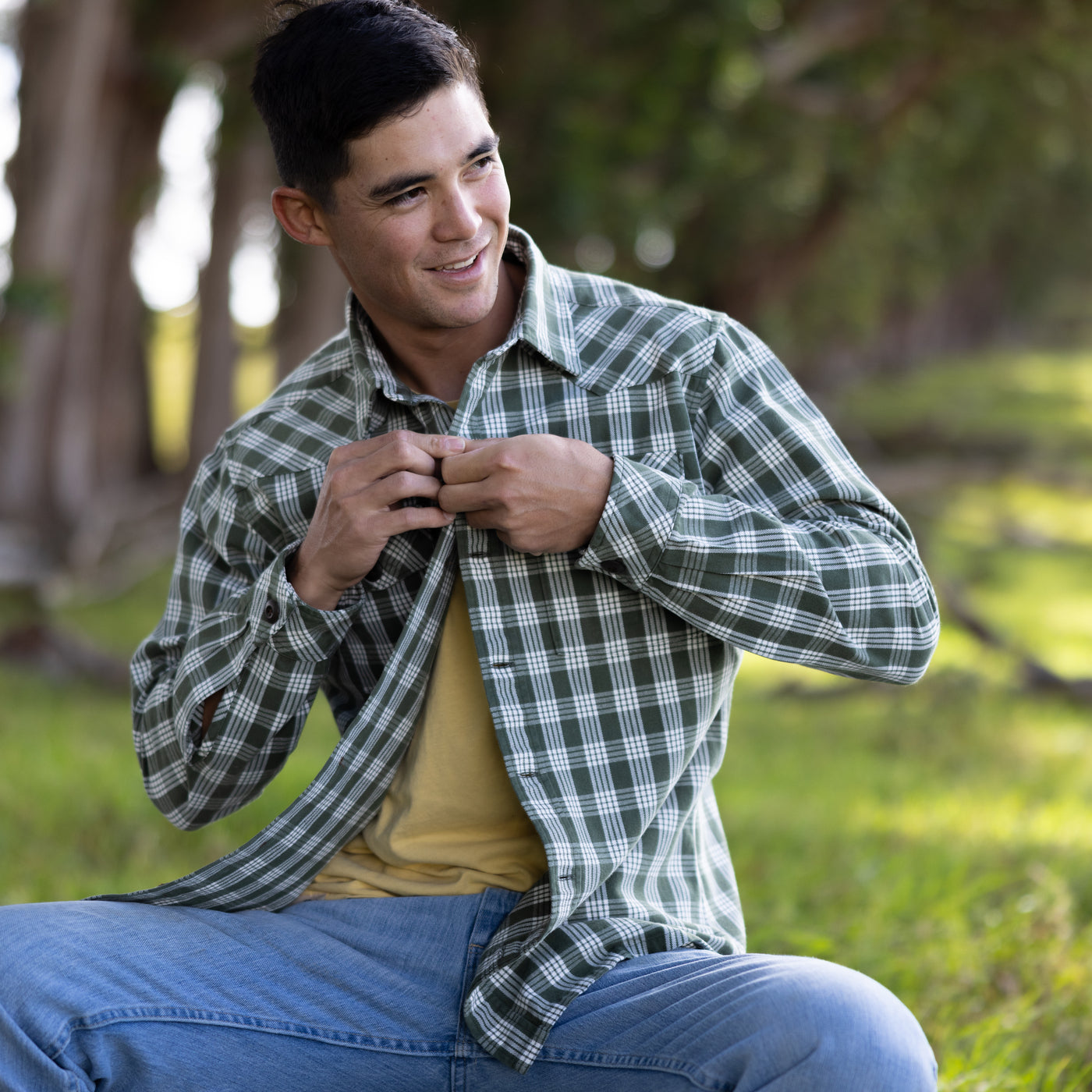 Man sitting outdoors wearing a green plaid shirt and blue jeans, smiling.