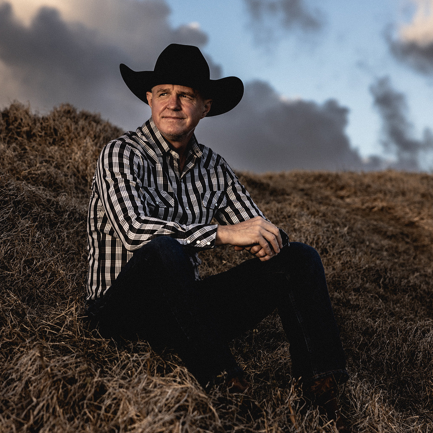 Man wearing a cowboy hat and checkered shirt sitting on a hay bale with a cloudy sky background