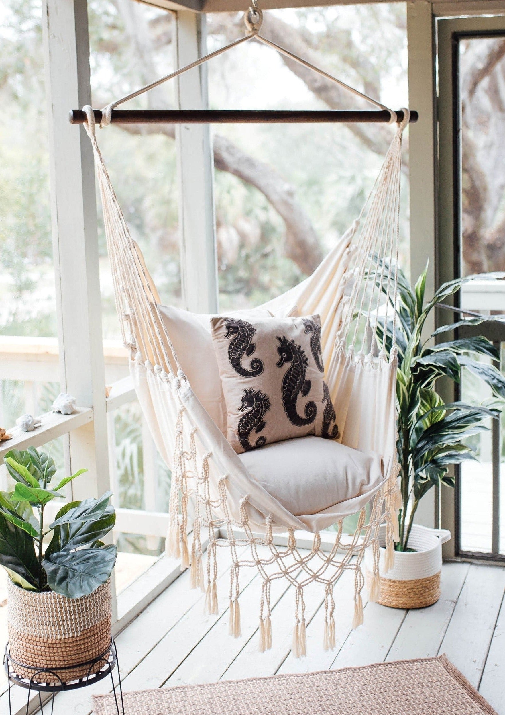 Beige hammock chair with decorative pillows in a bright room with large windows and plants.