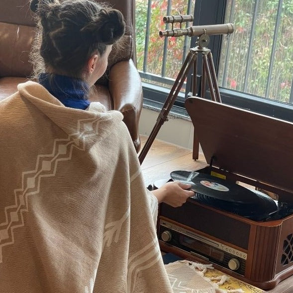 Person sitting on a couch with a vintage record player and vinyl record, wearing a cozy blanket and hat.