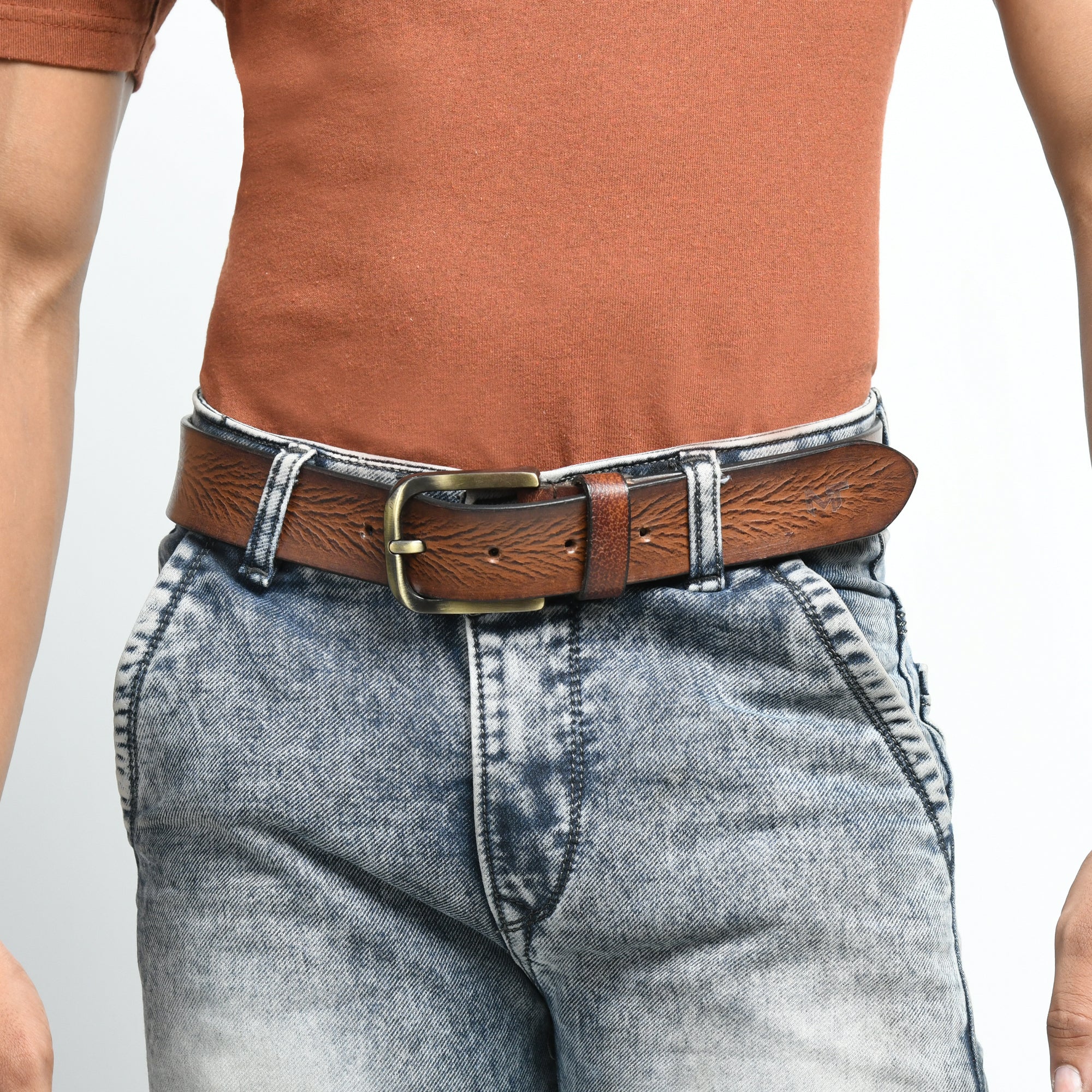 Person wearing a brown leather belt with a gold buckle on gray jeans against a white background