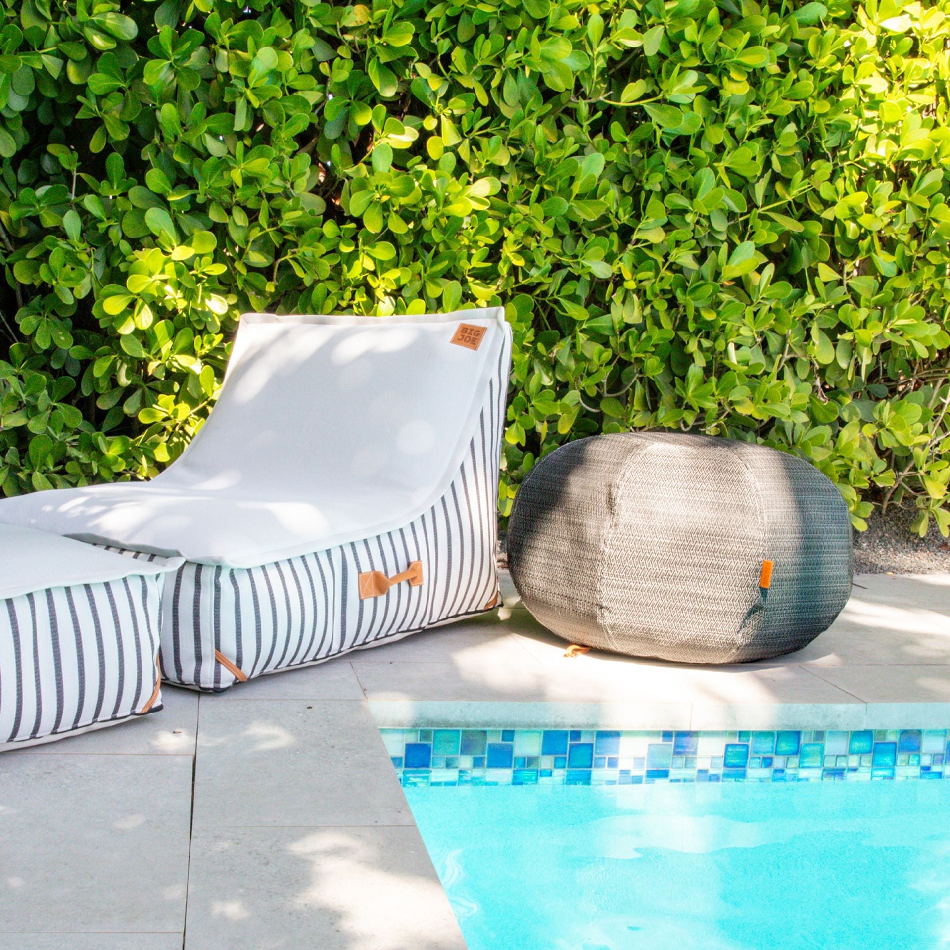Outdoor lounge chair and bean bag by a pool with greenery in the background