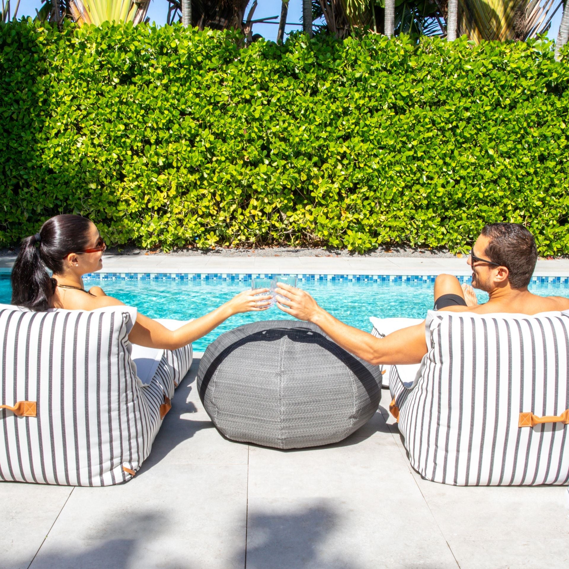 Two people sitting on striped bean bags by a pool with greenery in the background