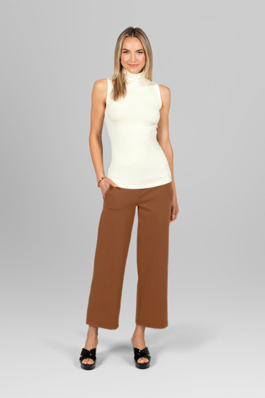 Woman wearing a white sleeveless top and brown pants on a gray background