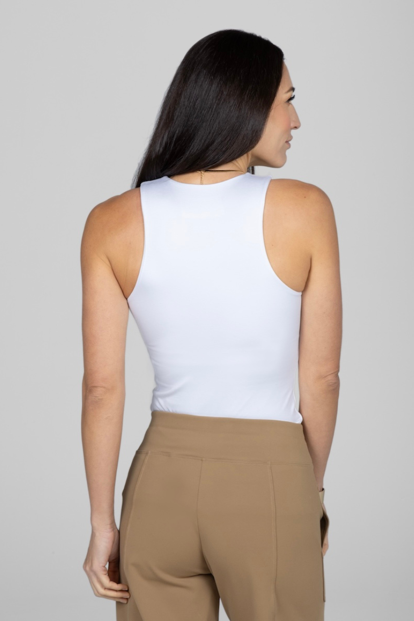 Woman wearing a white sleeveless top and beige pants against a plain background