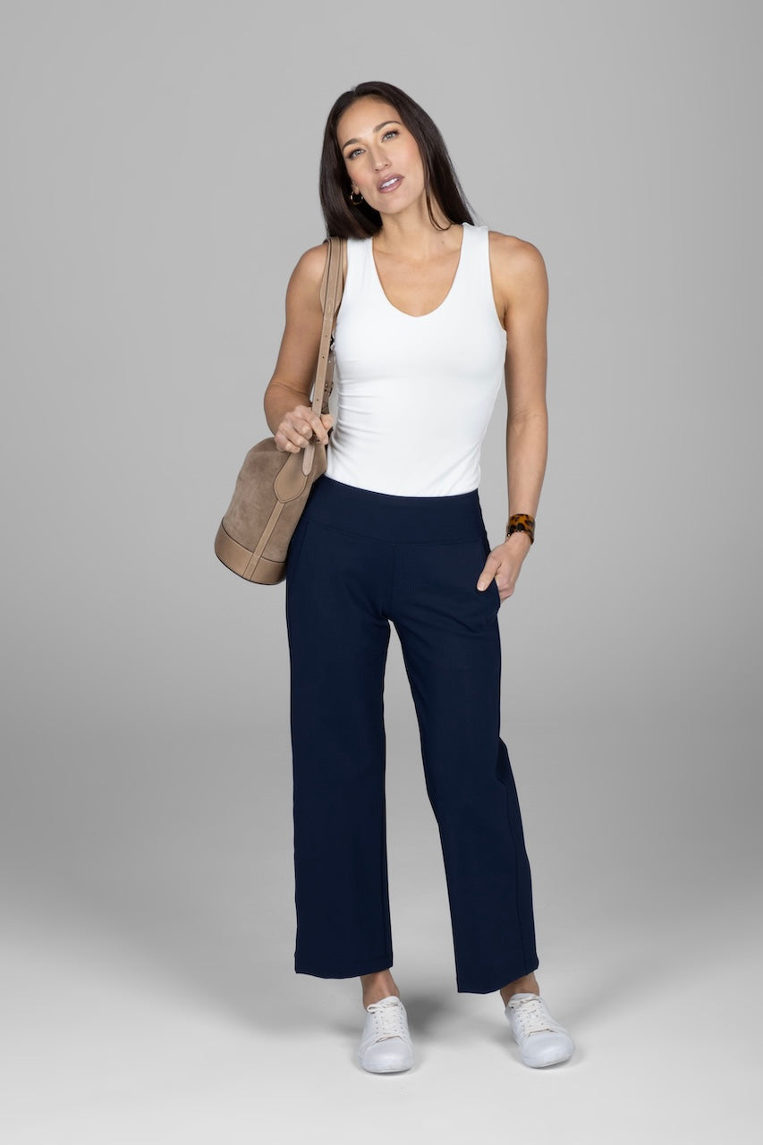 Woman wearing a white tank top and navy pants, holding a beige bag on a gray background