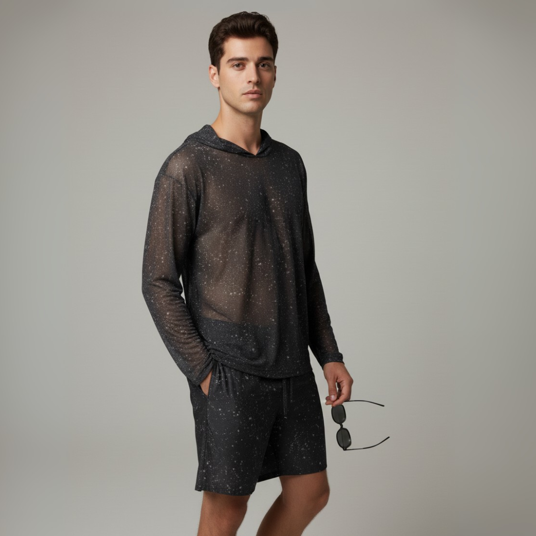 Man wearing a dark gray hoodie and shorts holding sunglasses on a plain background